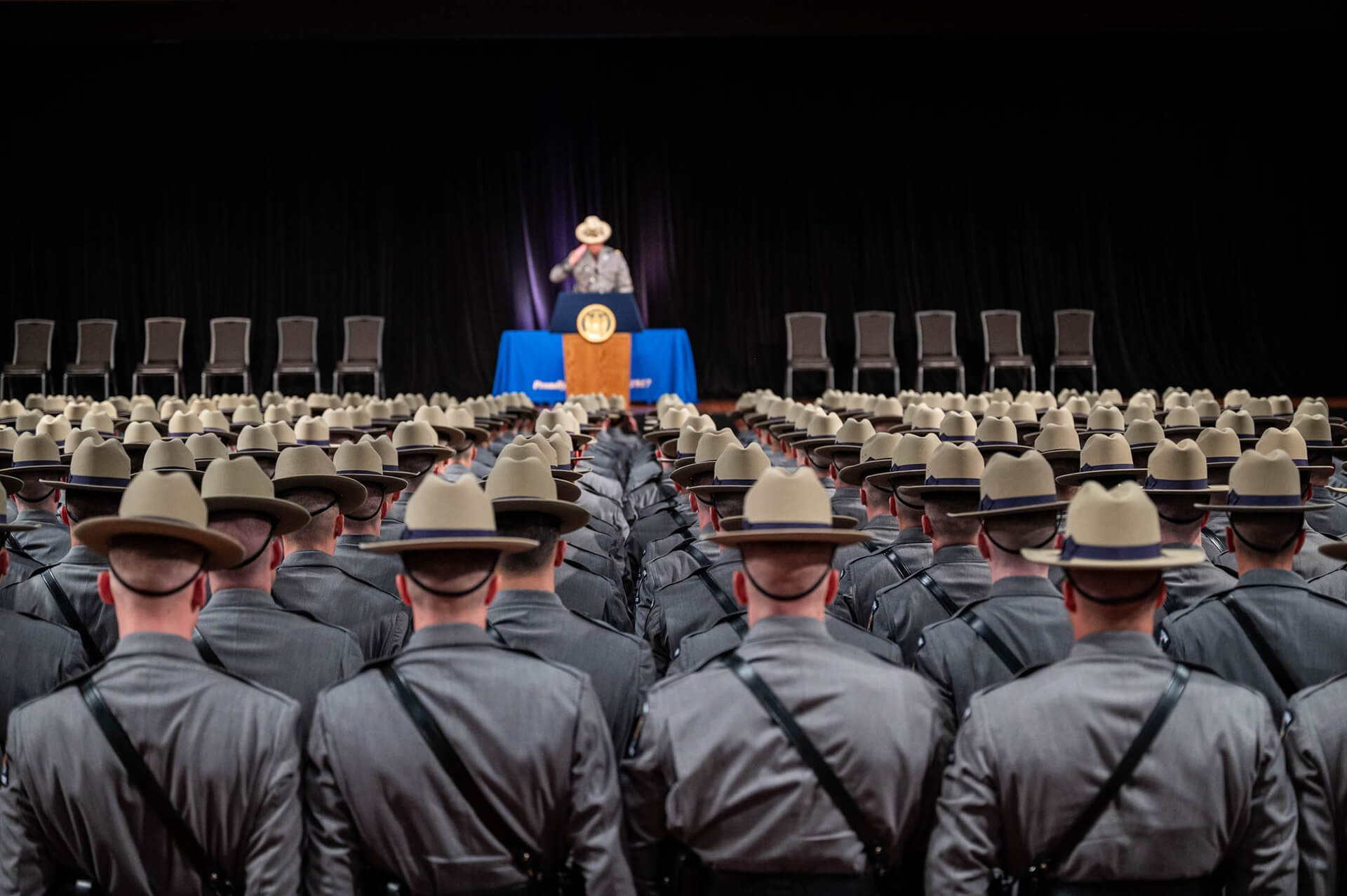 NY State Troopers standing in rows in front of a stage with an officer at the podium saluting